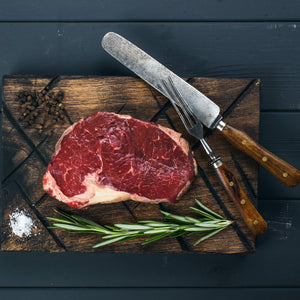 A beautiful grass-fed ribeye steak, perfectly marbled, sitting on a criss-crossed wooden chopping board. Salt, pepper and thyme sit beside it. A carving knife and fork are perched next to the steak. 